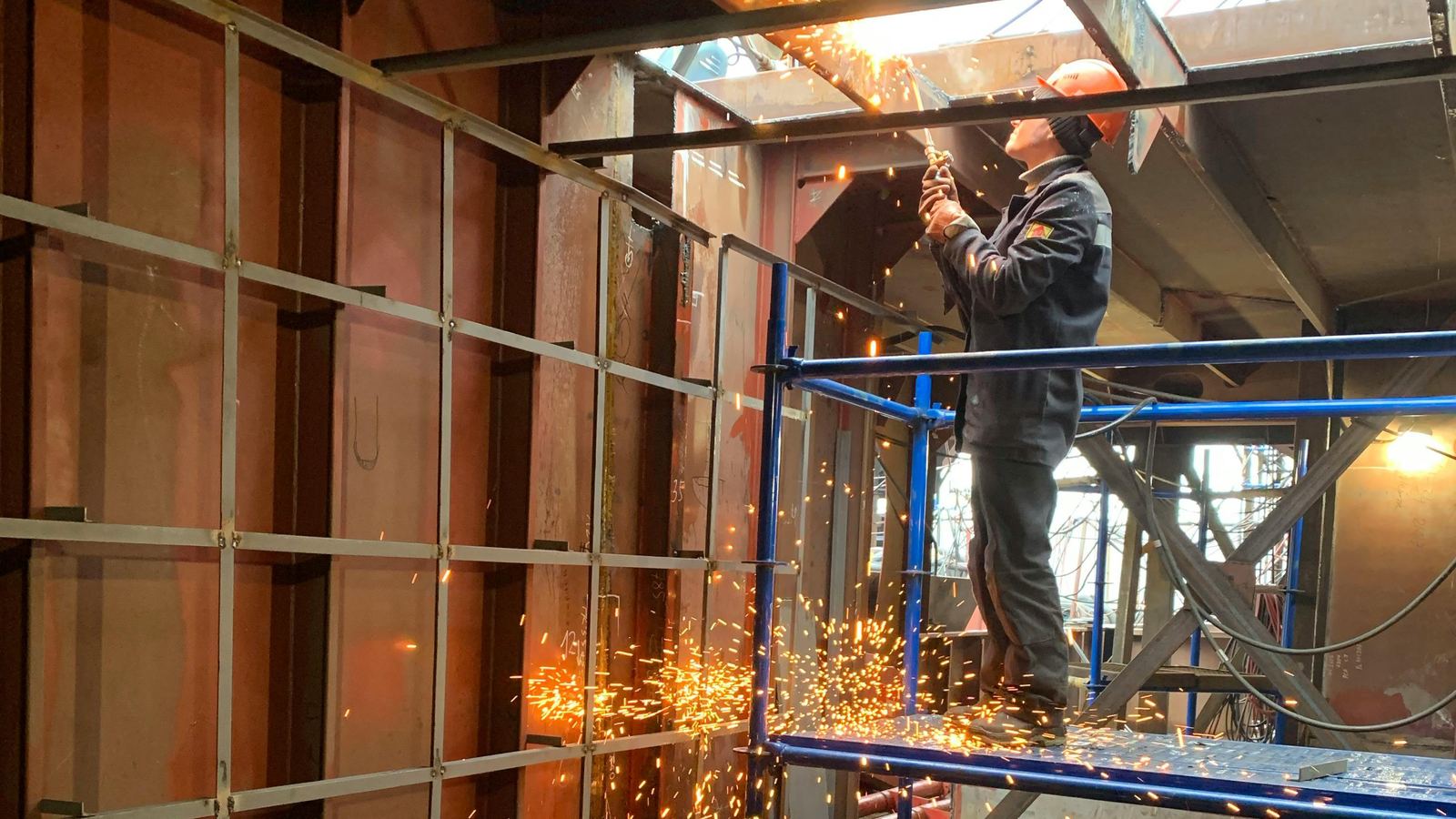 Welder on a scaffold working on the steel structure inside a ship's hull, with sparks falling