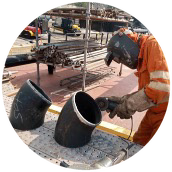 Welder in orange coveralls working on large-diameter pipe elbows