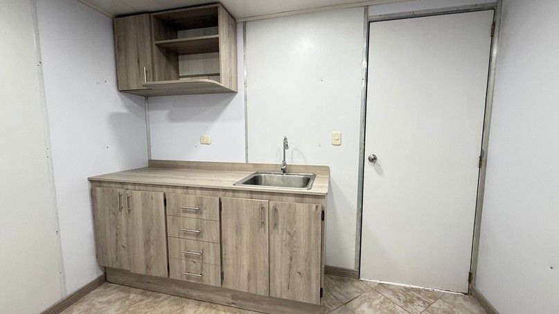 Kitchenette inside a trailer, with a sink, wooden cabinets and an upper shelf.