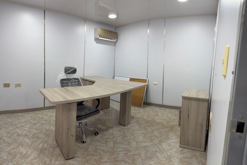 Interior of a trailer-type office with an L-shaped desk, a chair, a cabinet, a bulletin board and an air conditioner.