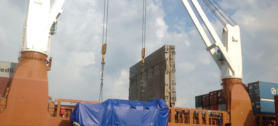 Port unloading of an engine covered by a blue tarp on a modular platform, with the ship’s cranes in the background.