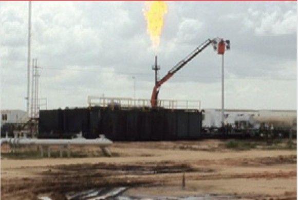 Field installation with a lit flare and a hydraulic arm next to a tank, in an operating area.