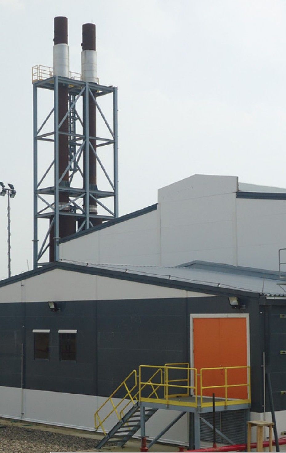 Machine hall building of a thermoelectric plant next to the exhaust stack tower, with access stairs and a white auxiliary unit.