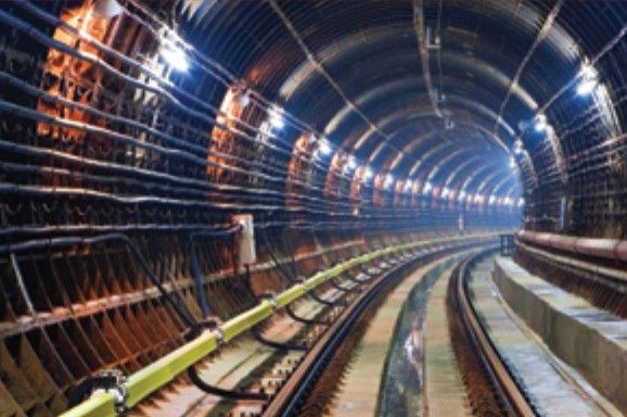 Inside a railway tunnel, with tracks, lighting and conduits along the wall.